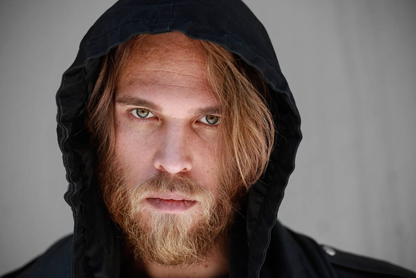 Headshot of brutal man with ginger thick beard, wears hoodie posing over black background with free space. - Foto, Imagem