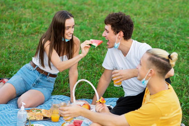 Grupo de amigos comendo e bebendo, se divertindo em um cobertor de piquenique com guloseimas, em um parque durante a pandemia em Chisinau, Moldávia - Foto, Imagem