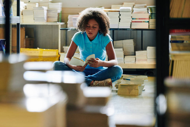Teenage girl sitting among book stacks in library and reading novel - Foto, Imagem