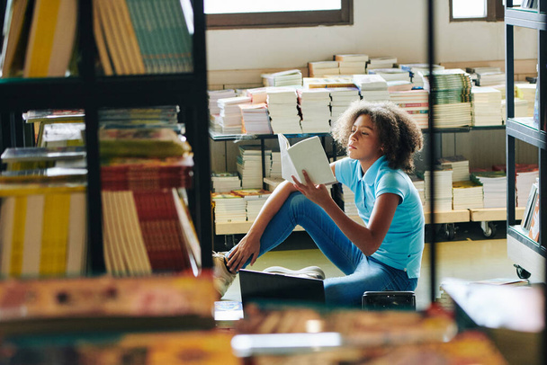Кучерява дівчина-підліток сидить на підлозі в бібліотеці і читає цікаву книгу
 - Фото, зображення