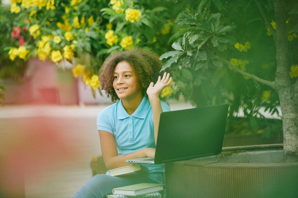 Досить усміхнена дівчина-підліток сидить на відкритому повітрі з ноутбуком і махає рукою
 - Фото, зображення