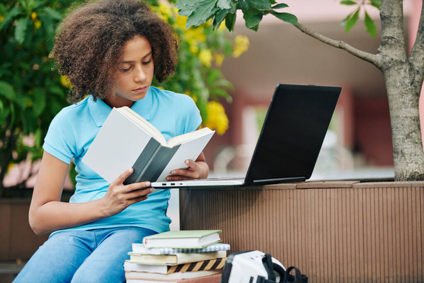 Kampüste ilginç kitaplar okuyan konsantre kıvırcık ırklı bir kız öğrenci. - Fotoğraf, Görsel