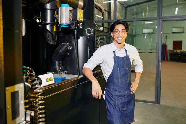 Portret van positieve barista in denim schort staan op grote moderne rooster en glimlachen op de camera - Foto, afbeelding