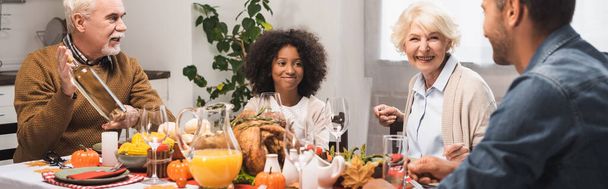 panoramic crop of senior man holding bottle of white wine during thanksgiving dinner with multicultural family - Photo, Image