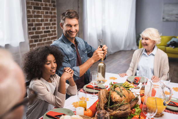 selective focus of man opening bottle of white wine during thanksgiving dinner with multicultural family - Foto, imagen