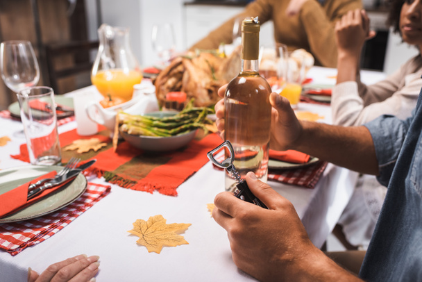 vista ritagliata dell'uomo in possesso di bottiglia di vino bianco e cavatappi durante la cena del Ringraziamento con famiglia multiculturale - Foto, immagini