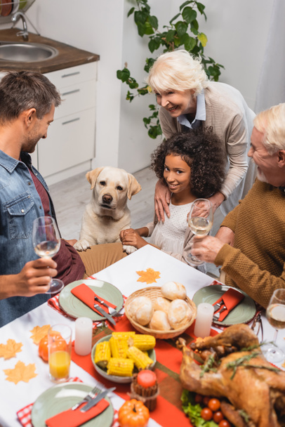 multicultural family and golden retriever near table served with festive thanksgiving dinner - Foto, Imagem