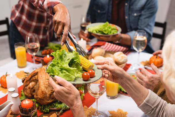 vista recortada de la mujer mayor sosteniendo plato con verduras durante la cena de acción de gracias con la familia multicultural - Foto, imagen
