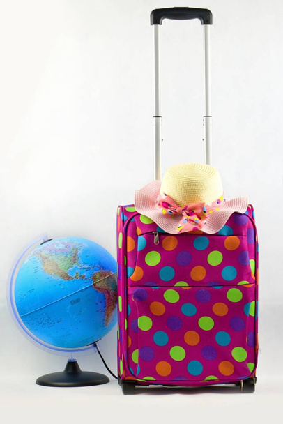 Rózsaszín bőrönd színes pöttyökkel, földgömbbel és női szalmakalapot elszigetelt fehér hátterben.Bőrönd - alapvető dolog az utazók számára. - Fotó, kép