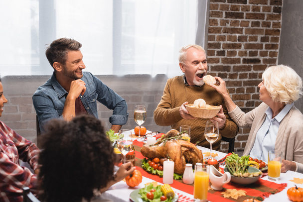 пожилая женщина кормит мужчину булочкой рядом с мультикультурной семьей во время ужина в честь Дня Благодарения - Фото, изображение