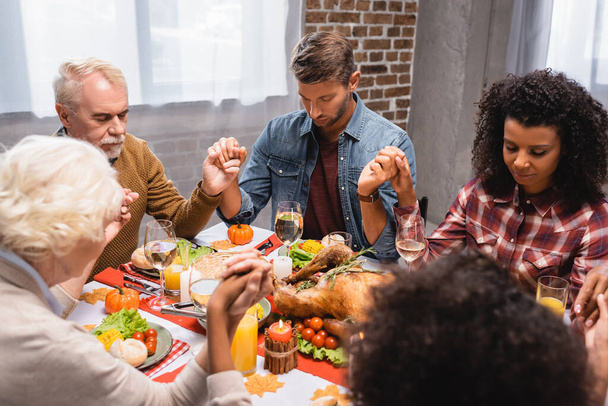 selective focus of multiethnic family with closed eyes holding hands during dinner on thanksgiving holiday - Foto, Imagen