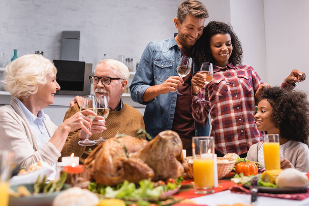 Foco seletivo da família multicultural com copos de vinho sentados à mesa com comida saborosa durante a ação de graças  - Foto, Imagem