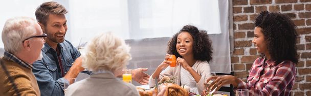 Panoramisch shot van multiculturele familie zitten in de buurt van smakelijke kalkoen op tafel tijdens Thanksgiving diner  - Foto, afbeelding