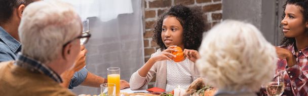 Panoramic orientation of african american girl holding pumpkin near parents and thanksgiving dinner  - 写真・画像