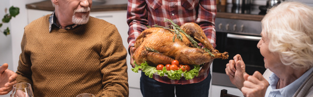 Panoramic shot of woman holding tasty turkey near family in kitchen  - Fotografie, Obrázek