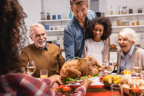 Selective focus of man looking at camera near multiethnic family and thanksgiving dinner with decor on table  - Фото, изображение