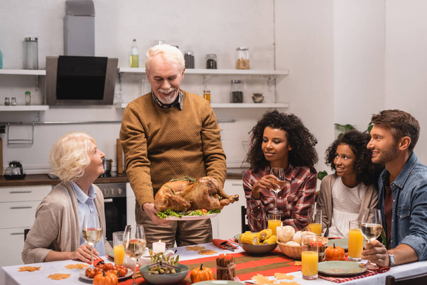 Selective focus of senior man holding delicious turkey near multicultural family   - Zdjęcie, obraz