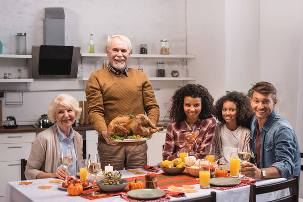 Selective focus of elderly man holding turkey near multiethnic family with wine during thanksgiving  - Photo, Image
