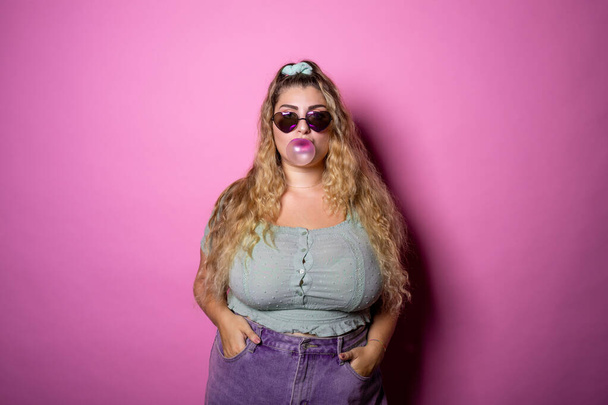Caucasian chubby young woman blows bubble from chewing gum isolated over pink background - Lovely diverse plus size woman chewing bubblegum - Positivity, charming, playful concept - Photo, Image
