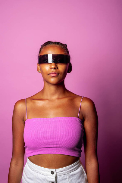 Junge schwarze Frau mit smarter Brille auf rosa Hintergrund - Diverse junge Frau mit Science-Fiction-Brille isoliert auf Hintergrund - Augmented Reality, werthaltige Technologie, Science-Fiction-Konzept - Foto, Bild