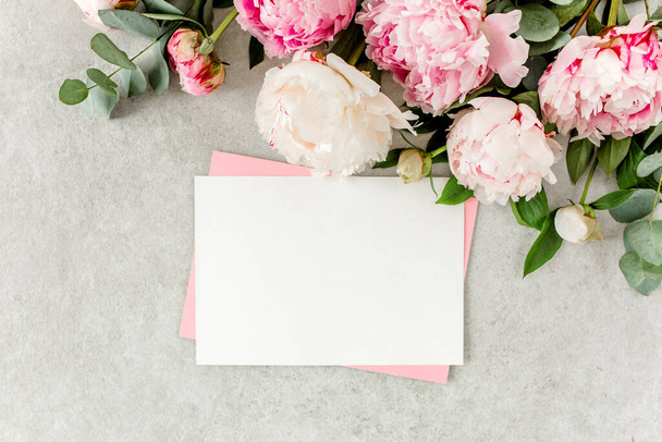 Mockup invitation, blank paper greeting card, pink envelope and peonies on gray stone table. Flower background. Flat lay, top view. - Фото, зображення