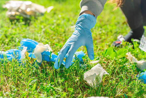 Coronavirus. Woman picking up blue rubber gloves and face masks during coronavirus epidemic lying on green grass. Environment contamination. Used. Garbage.  - Foto, Imagen