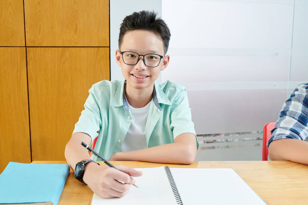Usmívající se vietnamský dospívající chlapec v brýlích psaní v copybooku a při pohledu na kameru - Fotografie, Obrázek