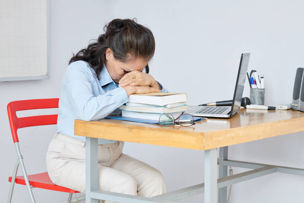 Uitgeput leraar leunend op stapel op studenten boeken op haar bureau om te rusten na een lange stressvolle dag - Foto, afbeelding