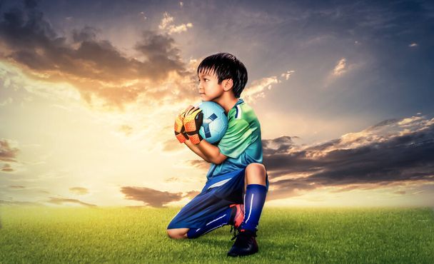 小さな子供のゴールキーパー成功サッカー劇的な概念のための黄金の空の背景と芝生のフィールドにボールを保持. - 写真・画像