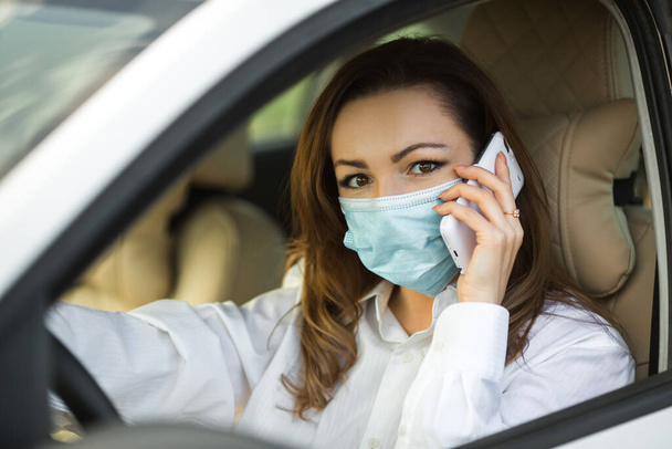 beautiful young woman in a medical mask driving a car with a mobile phone - Foto, Bild