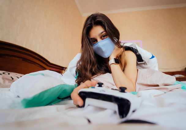 Dívka v ochranné masce ležící v posteli a držení ovladač - Fotografie, Obrázek