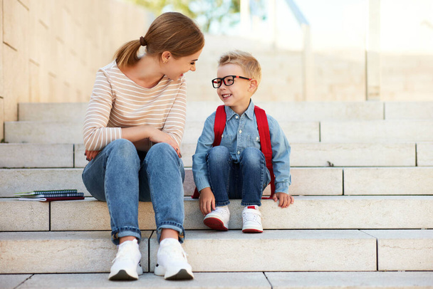  Anya és egy kisfiú, egy iskolásfiú könyvekkel és jegyzetfüzetekkel, együtt ülnek a lépcsőn az iskola első napján. - Fotó, kép
