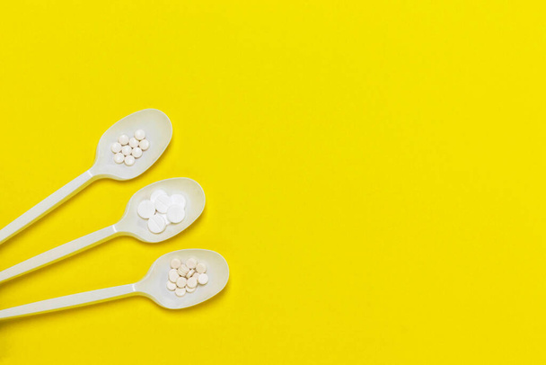 Three plastic spoons with pills on yellow background. Pharmaceutical industry. Epidemic, painkillers, healthcare and treatment concept. Flat lay. Top view with copy space. - Photo, Image