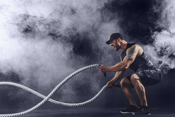 Bearded athletic looking bodybulder work out with battle rope on dark studio background with smoke. Strength and motivation - Foto, afbeelding