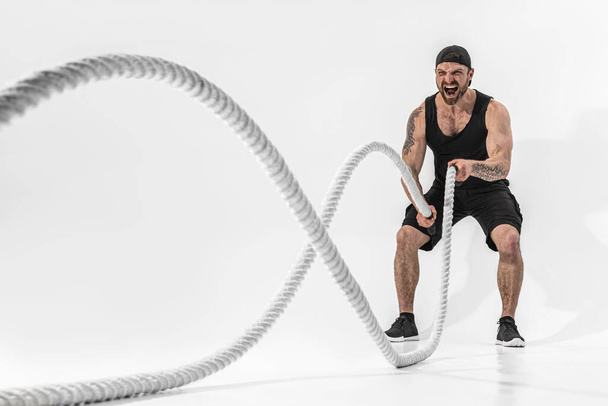 Attractive muscular man working out with heavy ropes. Photo of handsome man in sportswear isolated on white background - Zdjęcie, obraz