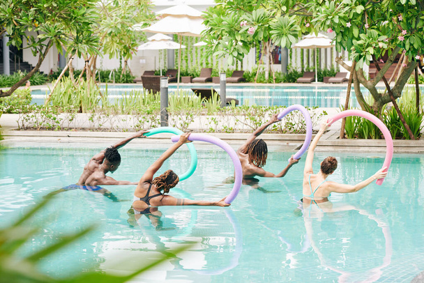 Група активних молодих людей, що стоять у басейні з плаваючою локшиною в руках, роблять бокові вигини, вид ззаду
 - Фото, зображення