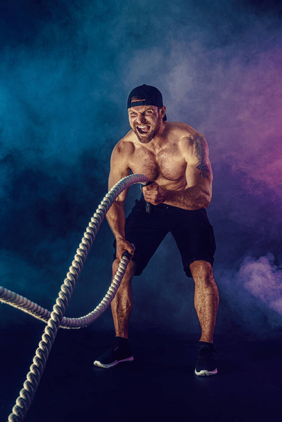 Bearded athletic looking bodybulder work out with battle rope on dark studio background with smoke. Strength and motivation - Foto, Imagem