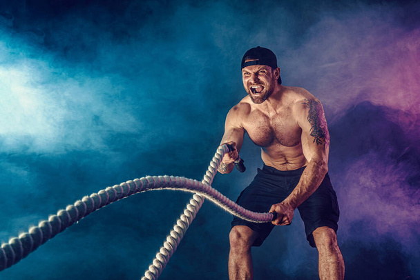 Bearded athletic looking bodybulder work out with battle rope on dark studio background with smoke. Strength and motivation - Foto, Bild