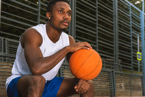 Jovem jogador de basquete afro-americano em camiseta branca e shorts azuis na quadra de basquete sentado, segurando basquete, concebida, conceito de esporte - Foto, Imagem