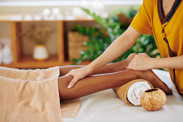 Imagen de cerca de la joven negra recibiendo masajes relajantes en las piernas en el salón de belleza y aplicando presión con el pulgar en el músculo de la pantorrilla - Foto, imagen