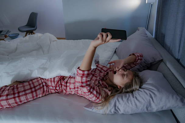  Bella giovane donna in pigiama che si estende a letto, spegne la sveglia su un telefono cellulare, avendo difficoltà a svegliarsi. - Foto, immagini