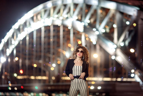 A girl in white clothes with black stripes stands against the background of the bridge. Night portrait of a girl in the style of the 90s. Model on the background of the night bridge. - Fotografie, Obrázek