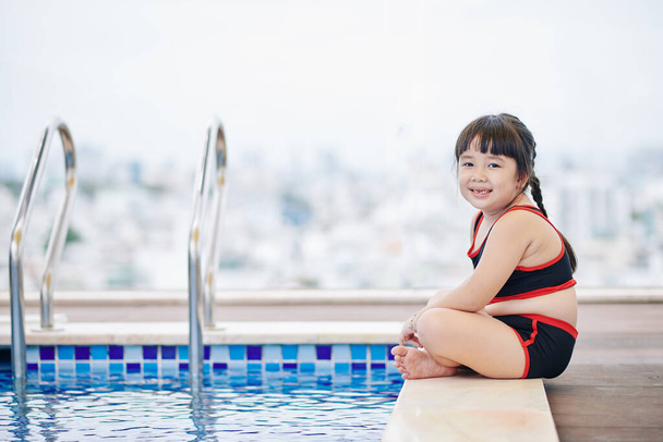 Χαρούμενη μικρή Βιετναμέζα που κάθεται στην άκρη της πισίνας και κοιτάζει την κάμερα. - Φωτογραφία, εικόνα