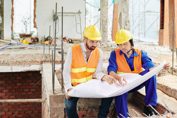 Serious multi-ethnic builders sitting in room of building under construction and discussing blueprint - Photo, Image