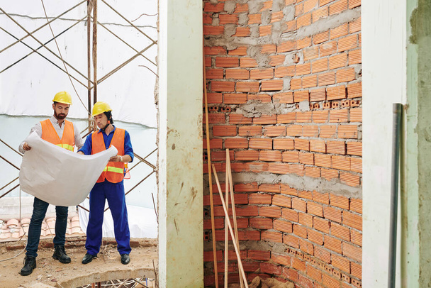 Trabajadores serios de la construcción de pie en la pared de ladrillo sin terminar y discutir plan de construcción - Foto, imagen