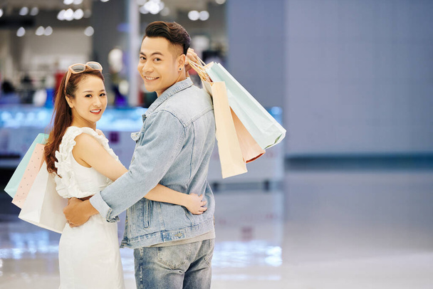 Portrait of cheerful young Asian man and women holding shopping bags, hugging and turning back to smile at camera - Foto, Imagen