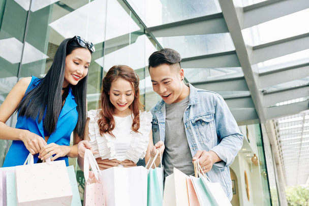Bastante sonriente joven vietnamita mostrando sus compras a amigos cuando están de pie en el centro comercial - Foto, imagen