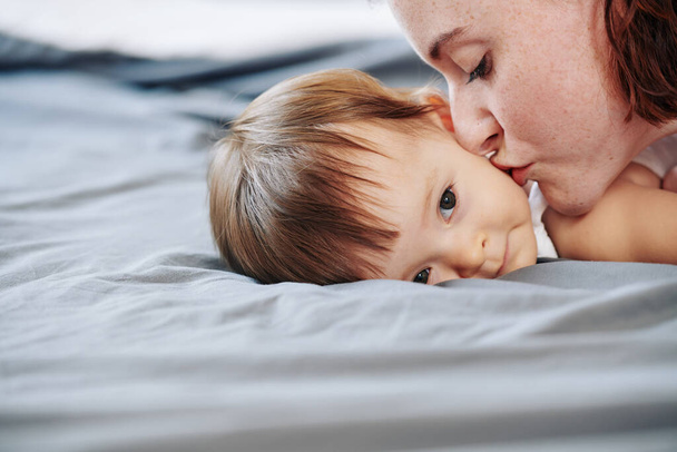 Immagine ravvicinata della giovane madre che bacia la sua piccola figlia sulla guancia quando riposa sul letto dopo il gioco - Foto, immagini