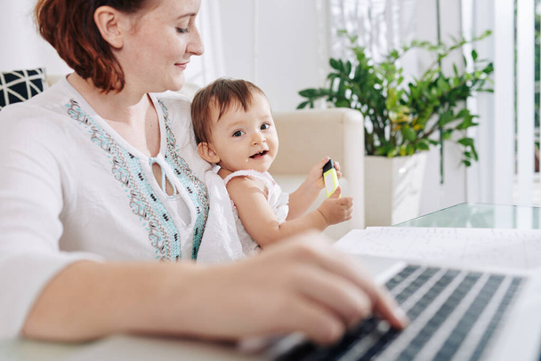 Linda niña feliz con resaltador en las manos sentado en las vueltas de su madre trabajando en el ordenador portátil - Foto, imagen