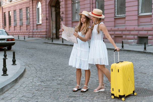 Twee meisjes zussen reizen. Het zijn toeristen en kijk naar de kaart in hun handen. Eén zus heeft een gele koffer bij zich.. - Foto, afbeelding